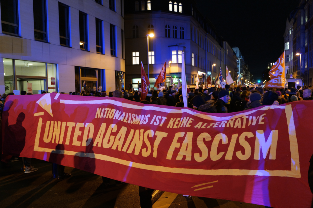 Eine Gruppe von Menschen marschiert mit einem Banner, auf dem 'Vereinigt gegen Faschismus' steht, eine Straße entlang, mit Gebäuden, Laternen und Schildern an der Straße und dem Himmel im Hintergrund.