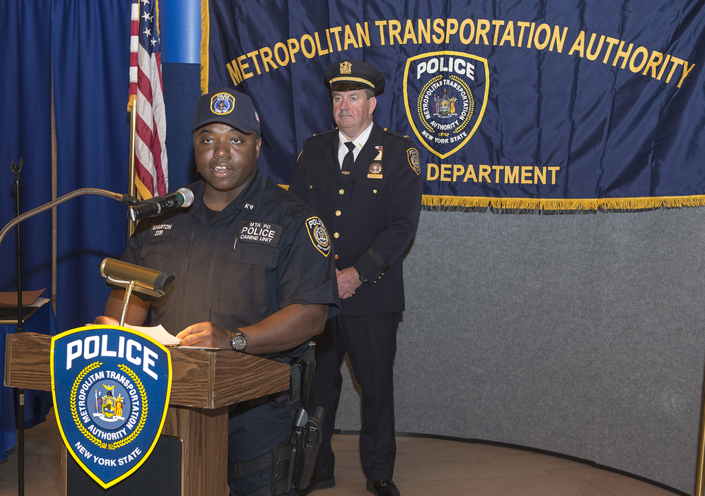 Zwei Polizisten stehen vor einem Podium, einer hält Papiere und spricht in ein Mikrofon, mit einer Tafel, Fahne und einem Tisch in der Nähe, und einer Tafel mit Symbolen und Text im Hintergrund.