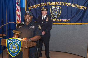 Zwei Polizisten stehen vor einem Podium, einer hält Papiere und spricht in ein Mikrofon, mit einer Tafel, Fahne und einem Tisch in der Nähe, und einer Tafel mit Symbolen und Text im Hintergrund.