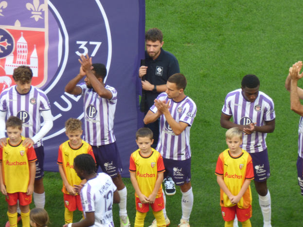 Eine Gruppe junger Jungs steht auf einem Fußballfeld, klatscht in die Hände, mit einem Banner und Logos im Hintergrund und saftigem Grün unter den Füßen.