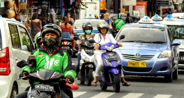 Eine Gruppe Motorradfahrer fährt auf einer vielbefahrenen Straße neben Autos und anderen Fahrzeugen, einige Fahrer tragen Helme, im Hintergrund sind Gebäude, Bäume, Schilder und Geschäfte zu sehen.