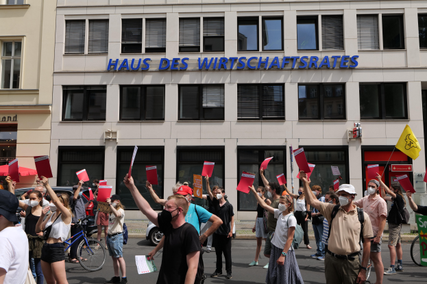 Eine Gruppe von Menschen, die vor einem Gebäude protestieren und Fahnen und Plakate halten, mit Fahrzeugen auf der Straße und einem Schild auf der rechten Seite.