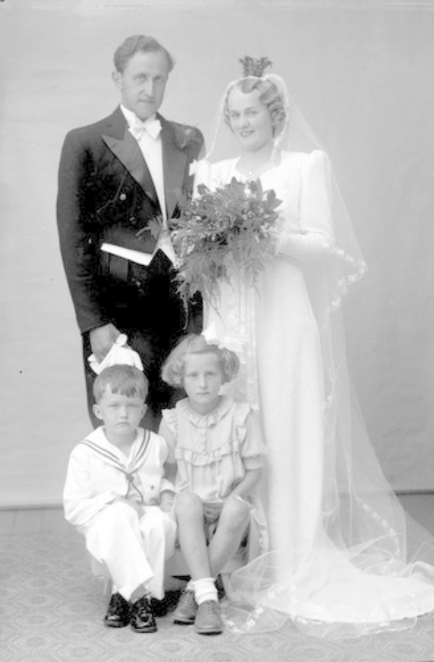 Schwarzes und weißes Foto einer Braut in einem weißen Kleid mit einem Strauß, Bräutigam lächelt neben ihr, und zwei Kinder sitzen vor ihnen auf dem Boden.