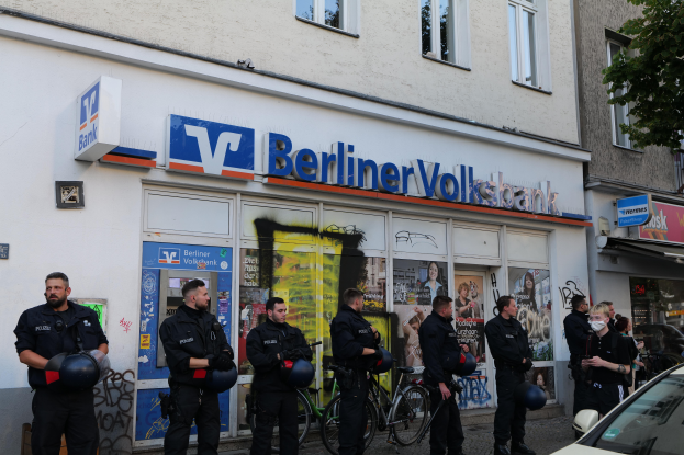 Eine Gruppe von Polizisten steht vor einer Berliner Volksbank, einige halten Helme in den Händen, mit einem Auto auf der rechten Seite, einem Baum auf der linken Seite und Gebäuden mit Fenstern und Texttafeln im Hintergrund.