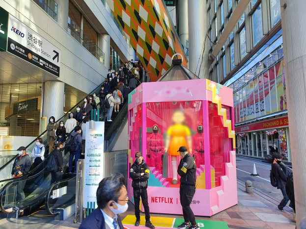 Eine Gruppe von Menschen steht vor einem Glaswand-Gebäude mit Texttafeln, einige tragen Masken, während eine Rolltreppe mit Menschen daneben fährt; in der Mitte eine pinkfarbene Box mit Schaufensterpuppen und der Aufschrift "Netflix's new Netflix Booth in Tokyo."