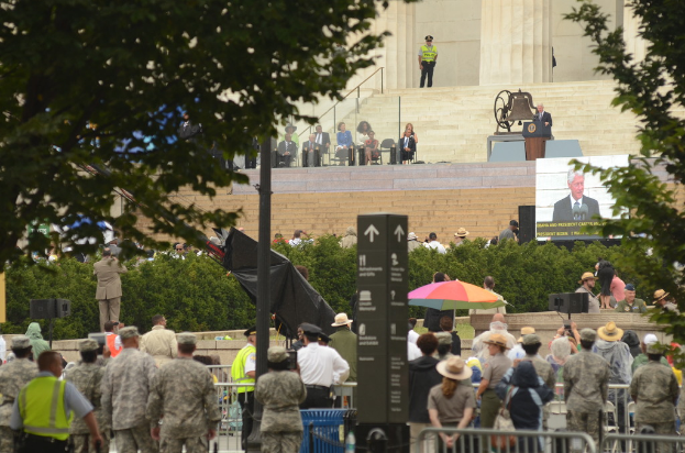 Army personnel and civilians stand and sit listening to a speech by the president, with trees, bushes, and stairs in the background.