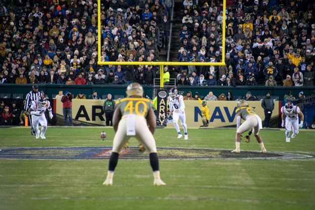 Fußballspieler in Helmen und Sportbekleidung konkurrieren auf einem Feld mit Zuschauern im Hintergrund, Stadioninfrastruktur einschließlich Pfosten und Tafeln mit sichtbarem Text.