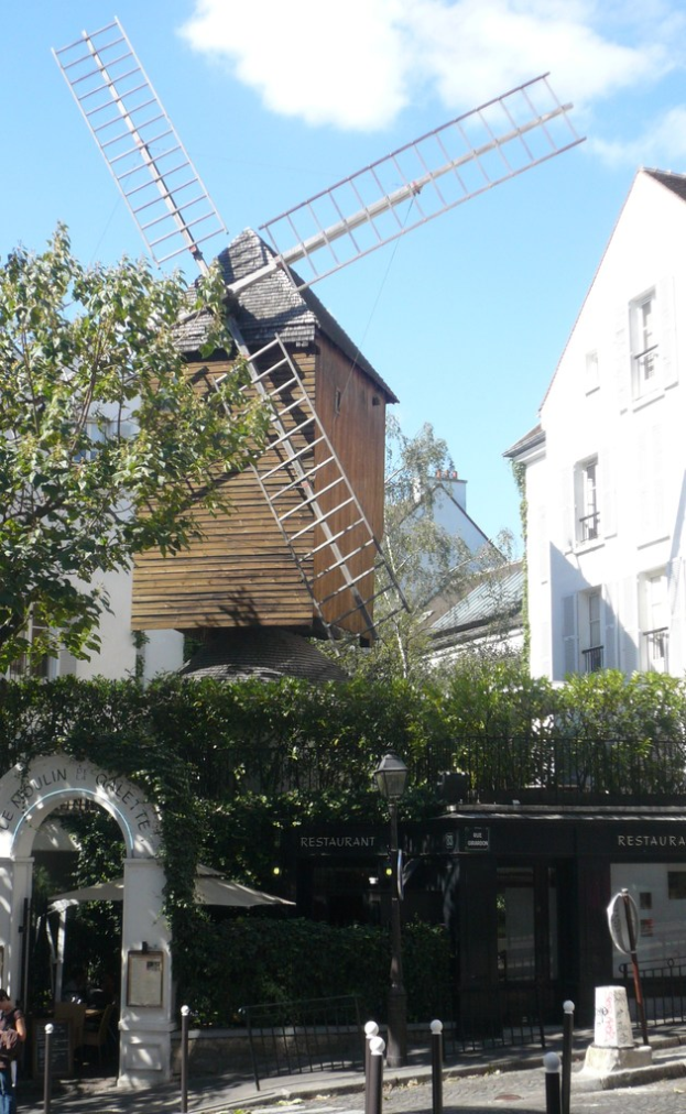 Eine Windmühle, mehrere Gebäude, Bäume, Metallstangen und ein Zaun.