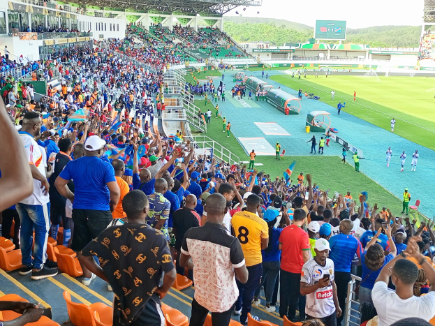 Eine große Menschenmenge versammelt sich vor einem eingezäunten Fußballfeld mit Hügeln und einem klaren blauen Himmel im Hintergrund, in Erwartung eines Spiels während der 2019 African Cup of Nations.