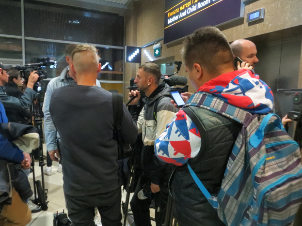 Eine Gruppe von Menschen, einige mit Kameras und Handys, andere mit Taschen, steht in einem Flughafen-Pressekonferenzbereich mit einem Textschild und Scheinwerfern dahinter.