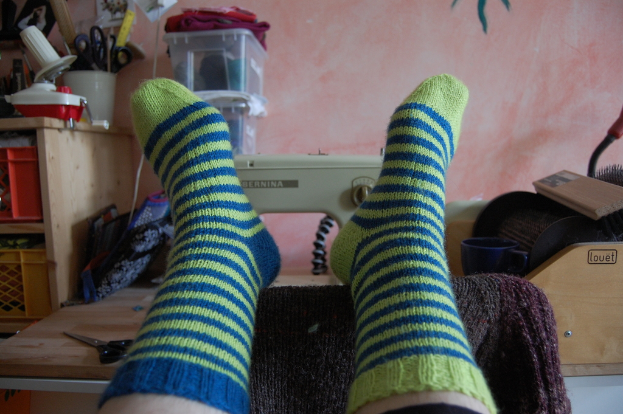 Eine Person in Socken steht neben einer Nähmaschine, Scheren, Boxen und Körben an einer Wand mit Kleidung.