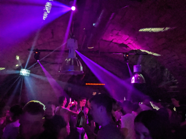 Eine Gruppe von Menschen, die in einem Nachtclub tanzen, unter lila und blauen Deckenlichtern, einige halten Flaschen.