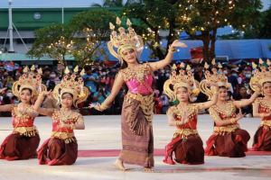 Eine Gruppe von Frauen in traditioneller Thai-Tracht, die vor einem Publikum tanzen, mit Bäumen, Lichtern und einem Gebäude im Hintergrund unter einem klaren blauen Himmel.
