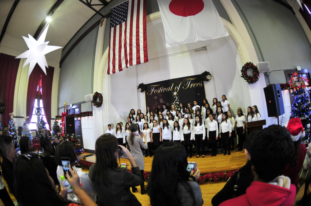 Eine Gruppe von Menschen auf einer Bühne vor einem Publikum, mit geschmückten Weihnachtsbäumen, einem Rednerpult, Lautsprechern, einer Texttafel, Vorhängen, Flaggen und Deckenleuchten.