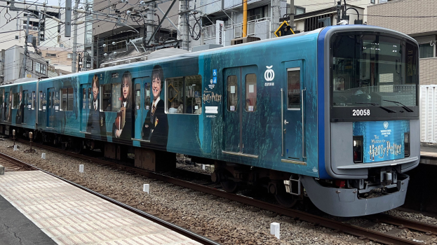 Blauer Zug fährt auf Schienen neben einem Bahnsteig mit elektrischen Masten und Drähten, Gebäuden und wolkenlosem Himmel im Hintergrund.