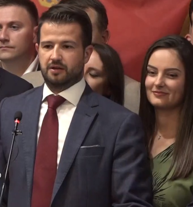 Ein Mann im Anzug spricht an einem Rednerpult mit einem Mikrofon zu einer Menge hinter einer Fahne, wahrscheinlich über ein Regierungsozialmedienverbot.