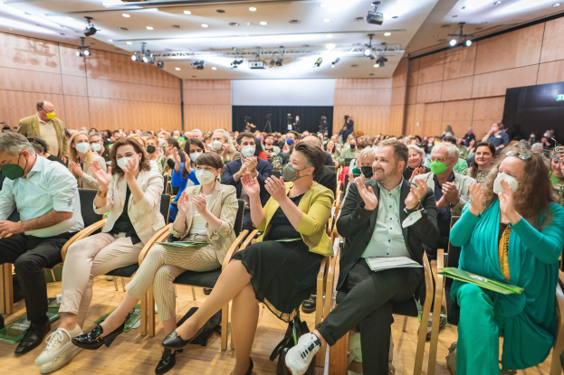 Eine Gruppe von Menschen in Stühlen klatschend, einige tragen Masken, mit Taschen auf dem Boden, vor einer Menge bei einer Coronavirus-Konferenz mit einem Bildschirm und Deckenbeleuchtung im Hintergrund.