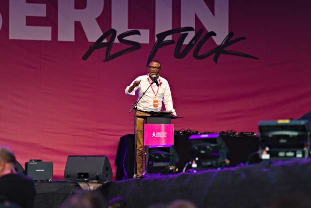 Ein Mann in einem weißen Hemd und braunen Hosen steht auf einer Bühne und hält ein Mikrofon, während ein pinker Banner mit der Aufschrift 'Berlin as Fuck' hinter ihm und Lautsprecher auf der Bühne zu sehen sind.