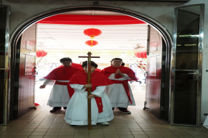 Menschen, die mit einem Kreuz in einer Kirche gehen, mit Türen links und rechts und Blumen und dekorativen Elementen im Hintergrund.