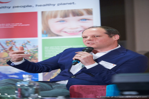 Ein Mann in einem schwarzen Anzug sitzt auf einem Stuhl und hält ein Mikrofon, mit einem Bannerboard dahinter.