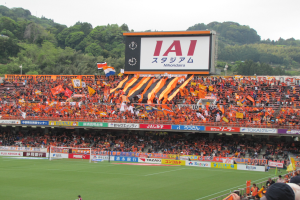 Ein Fussballspiel wird in einem Stadion mit einer grossen Zuschauermenge, grünem Rasen, einem Tor, Bannern, Fahnen, einem grossen Bildschirm, Bäumen und einem klaren blauen Himmel gespielt.