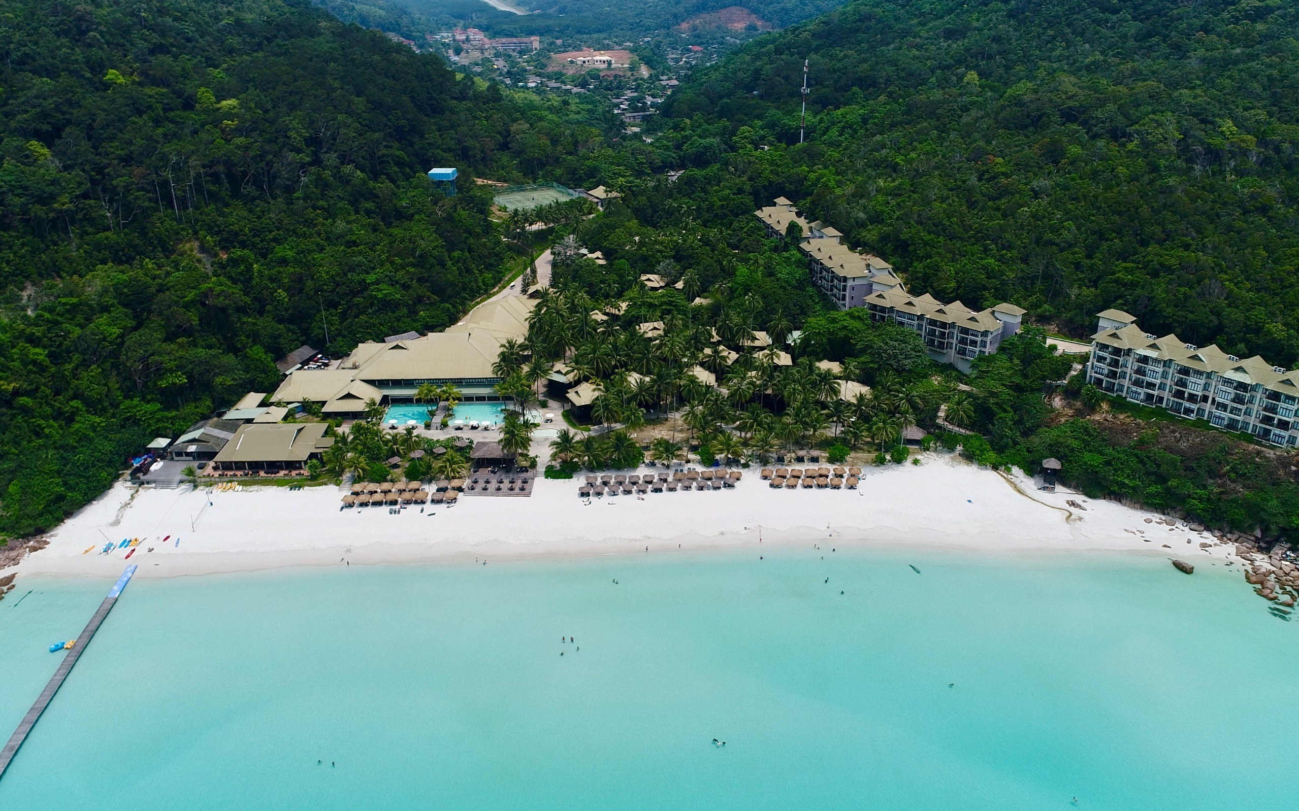 Luftaufnahme eines Strandresorts in Koh Samui, Thailand, mit einer Brücke links, Gebäuden, Bäumen, Hügeln und Menschen am Strand.