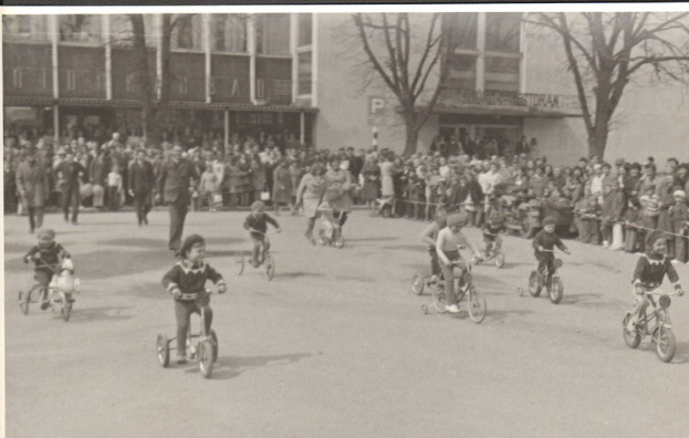 Schwarzes und weißes Bild von Kindern, die auf Fahrrädern eine Straße entlangfahren, mit Zuschauern am Straßenrand, Bäumen und Gebäuden im Hintergrund und Text unten.