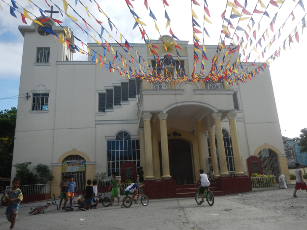 Eine Gruppe von Menschen, die Fahrräder vor einer weißen Kirche fahren, mit architektonischen Details wie Fenstern, Säulen, Bögen, Türen und einem Kreuzsymbol, sowie Pflanzen, einem Zaun, einer Fahne, Bäumen und einem bewölkten Himmel im Hintergrund, und bunten Fahnen, die an einem Seil befestigt sind.