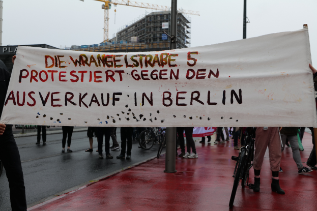 Eine Gruppe von Menschen steht auf der Stra├če und h├Ąlt ein Schild mit der Aufschrift "Die Wrangellstra├če 5 Protestiert Gegen den Ausverkauf in Berlin". Es sind Fahrr├Ąder, Pfosten, Geb├Ąude und ein Kran im Hintergrund zu sehen, ├╝ber ihnen der Himmel.