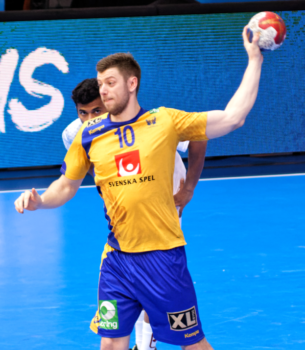 Ein Mann in einem gelben Hemd und blauen Shorts spielt Handball auf einem Platz, hält einen Ball und hat im Hintergrund einen Bildschirm mit der Aufschrift "Svenský Spel Handball-Weltmeisterschaft 2019".
