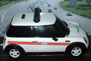 Ein weißer Polizeiwagen steht im Vordergrund mit mehreren fahrenden Autos auf der Straße dahinter, umgeben von Gras und Bäumen auf beiden Seiten.