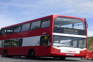 Ein roter Doppeldeckerbus mit der Aufschrift "Stadtbus" fährt auf der Straße, vor ihm steht ein Verkehrskegel und im Hintergrund ist ein Hügel zu sehen.