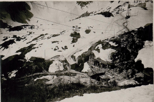 Ein Schwarz-Weiß-Foto eines Skilifts, der eine schneebedeckte Bergkulisse hinauffährt, mit Häusern, Pfählen und Drähten im Vordergrund und Text am unteren Rand.