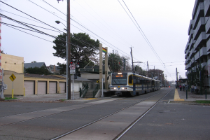 Eine Stadtbahn fährt durch eine Straße mit hohen Gebäuden, Strommasten und einigen Fußgängern, mit Bäumen und einem klaren blauen Himmel im Hintergrund.