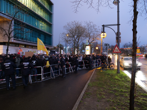 Eine große Gruppe von Menschen steht vor einem Gebäude in Berlin und hält Protestschilder mit Barrikaden, Fahrrädern, Schildern, Bäumen und Gras im Vordergrund.