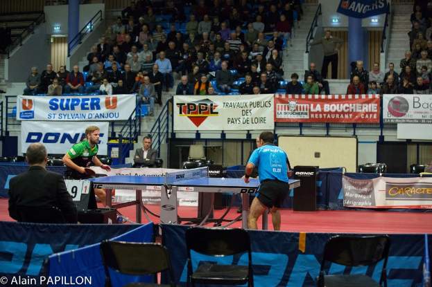 Zwei Männer spielen Tischtennis drinnen, mit mehreren Zuschauern, die stehen und sitzen, einigen leeren Stühlen und Werbeplakaten, mit Text in der linken unteren Ecke.