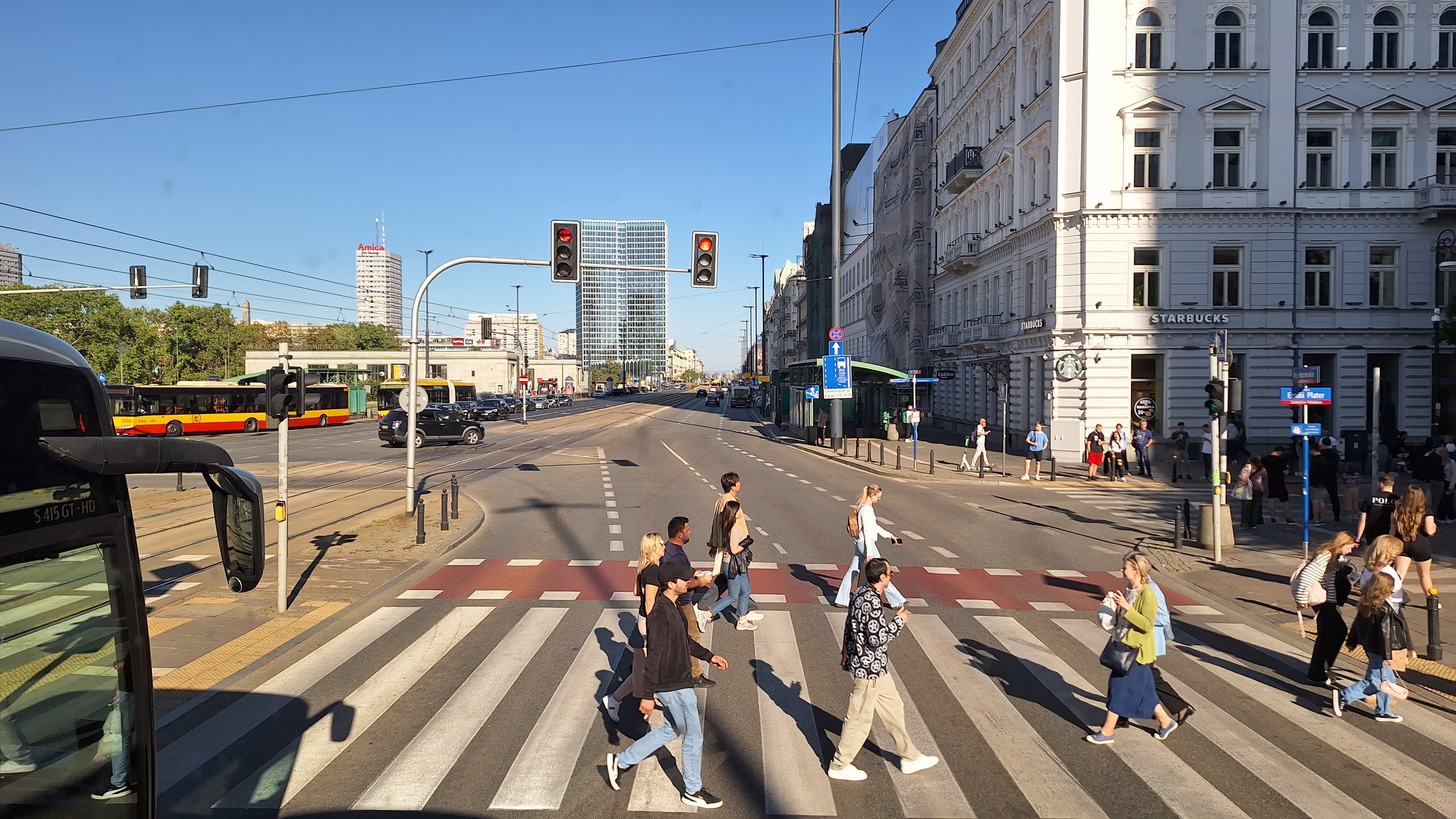 Gruppe von Menschen, die eine Straße an einer Ampel überqueren, mit einem Bus auf der linken Seite, Fahrzeugen auf der Straße, Verkehrsampeln, Laternenmasten, Gebäuden, Bäumen und einem klaren blauen Himmel im Hintergrund.