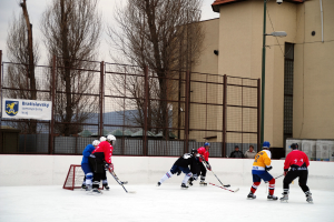 Personen beim Eishockeyspielen auf einem Eis mit Gebäuden, Bäumen, einer Straßenlaterne, einem Namensschild und Zäunen im Hintergrund unter einem Himmel.