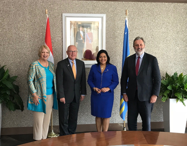 Eine Gruppe von Menschen, die um einen Tisch mit Papieren stehen und lächeln, umgeben von Fahnen, Topfpflanzen und einem Rahmenfoto an der Wand, was die Unterzeichnung eines U.S.-Canada-Memorandums of Understanding anzeigt.