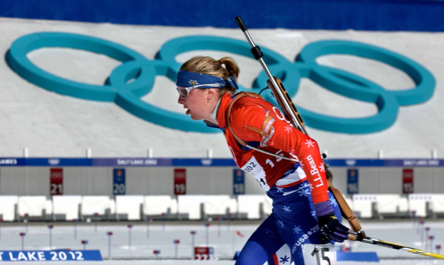 Lindsey Vonn in einem roten und blauen Skioutfit fährt eine schneebedeckte Piste hinunter und hält Ski-Stöcke in den Händen, während im Hintergrund Schilder mit Text und Zahlen sowie eine gestaltete Wand zu sehen sind.
