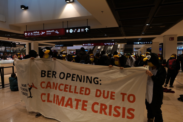 Gruppe von Menschen in einem Flughafen, die Masken tragen und ein Banner halten, auf dem "Ber Opening Cancelled Due to Climate Crisis" steht, einige stehen und andere sitzen auf dem Boden, Rolltreppen und Abflugtafeln sind im Hintergrund zu sehen.