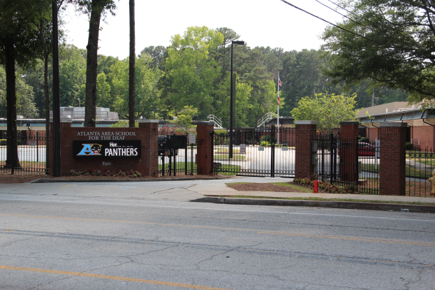 Eingang einer Schule mit einem Schild, umgeben von Bäumen, Straßenmöbeln, Fahrzeugen und Gebäuden unter einem klaren blauen Himmel.