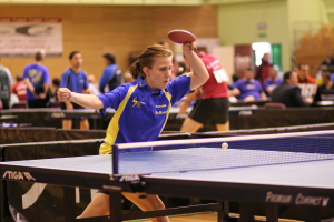 Eine Frau mit einem Schläger in der Hand wird hervorgehoben und spielt Tischtennis, umgeben von ein paar sitzenden und stehenden Personen und einem Banner an der Wand.