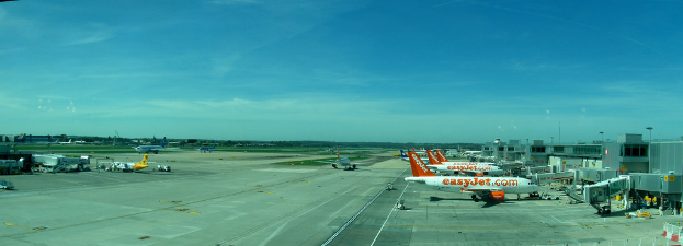 Eine Flughafen-Szene mit Flugzeugen, Gebäuden, Masten, Fahrzeugen und Bäumen, mit einem Himmel darüber und einer Straße darunter und einem Zaun im Vordergrund.