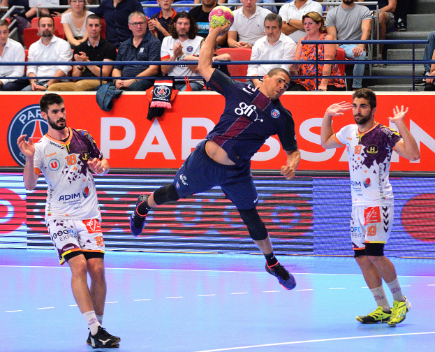 Eine Gruppe von Männern, die Handball auf einem Platz mit einem Ball in der Mitte spielen, mit Zuschauern im Hintergrund und verstreuten Kleidungsstücken auf dem Platz, mit einem Banner, auf dem "Futsal-Weltmeisterschaft 2015 - Paris Saint-Germain vs. Olympique Lyon" steht.