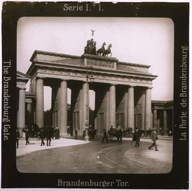 Ein Schwarz-Weiß-Foto des Brandenburger Tors in Berlin, Deutschland, mit einem zentralen Torbogen mit Säulen und Statuen, Menschen, die davor gehen, Bäumen auf der rechten Seite und Text unten.