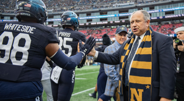 Ein Mann im Anzug und Krawatte gibt einem Fußballspieler die Hand auf einem Feld, umgeben von einer Gruppe von Menschen, mit einem Kameramann auf der rechten Seite und einem Stadion voller Zuschauer im Hintergrund, auf dem Schilder stehen, die "Notre Dame Football Coach Mike McCarthy und die Fighting Irish" zeigen.