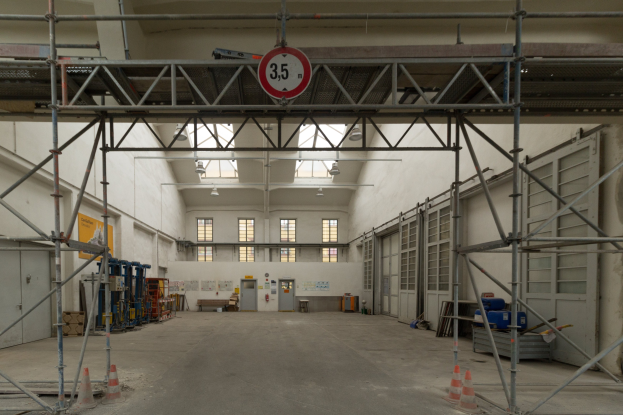 Lagerhalle mit ausgedehnten Gerüsten, Verkehrskegeln, Metallstangen, einem Schild, Maschinen, einer Tür, Fenstern, Wänden, Deckenlampen und Dach.