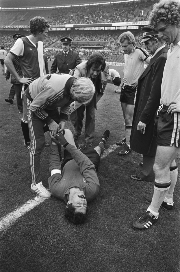 Schwarzes und weißes Foto eines Fußballspielers, der auf dem Boden liegt, während ihn eine Gruppe von Männern und eine Stadionmenge umgibt.