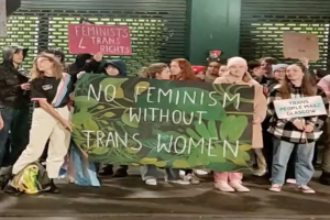 Gruppe von Menschen mit einem Banner, das "Kein Feminismus ohne Trans-Frauen" und Schilder in einer öffentlichen Fläche mit einer Wand und einem Rollladen im Hintergrund zeigt.